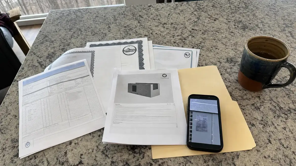 Equipment certification paperwork and model tag photographs spread out for a Georgia heat pump rebate claim on a mountain kitchen counter